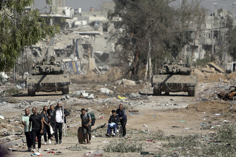 People walk along a road with tanks behind them pointing in their direction. Destroyed buildings seen in the backgroung