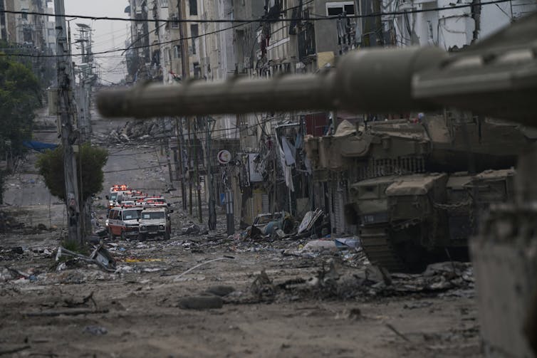 A tank on a narrow street with damaged buildings. Ambulances are seen in the background.