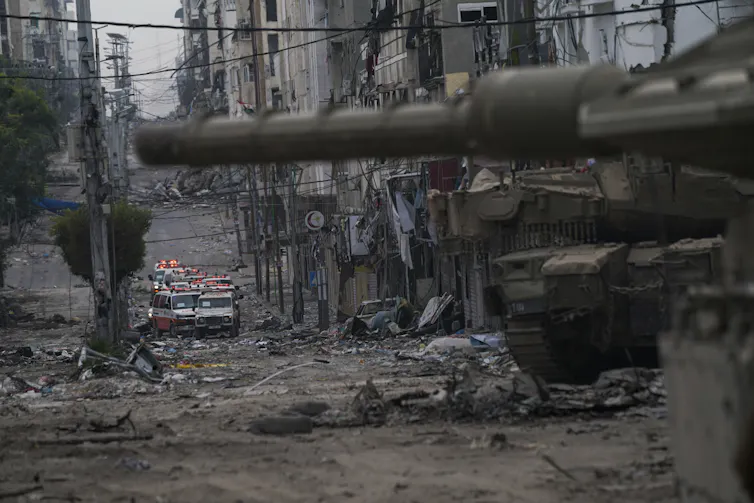 A tank on a narrow street with damaged buildings. Ambulances are seen in the background.