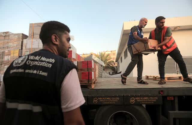 Two men carry boxes off a truck, a third man stands to  the side. He wears a vest from the World Health organization