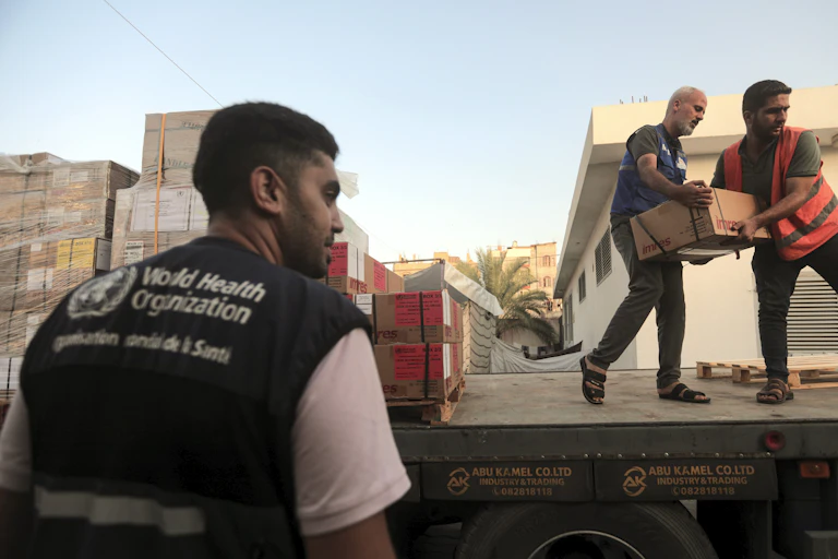 Two men carry boxes off a truck, a third man stands to the side. He wears a vest from the World Health organization