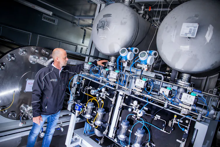 A man points to a wall of machinery and tanks.