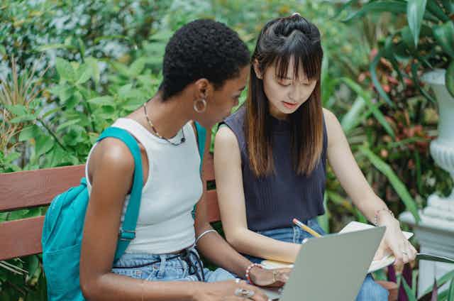 Two young women sit on a bench with a laptop and a notebook.