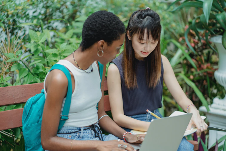 Two young women sit on a bench with a laptop and a notebook.