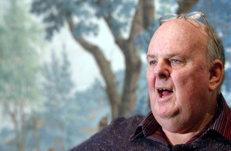 A head shot of poet Les Murray against the backdrop of a landscape painting.