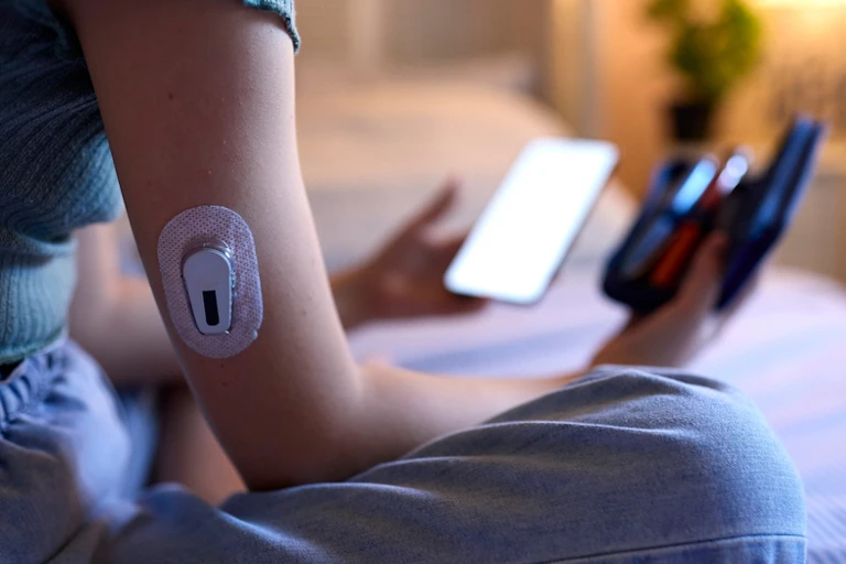 Teenage girl sitting on bed with glucose monitor patch on arm, holding smartphone and receiver