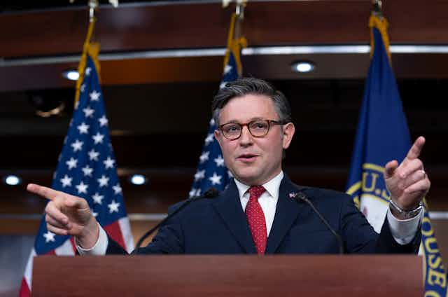 A man wearing a navy jacket and glasses speaks from a lectern with the flags of the United States behind him.