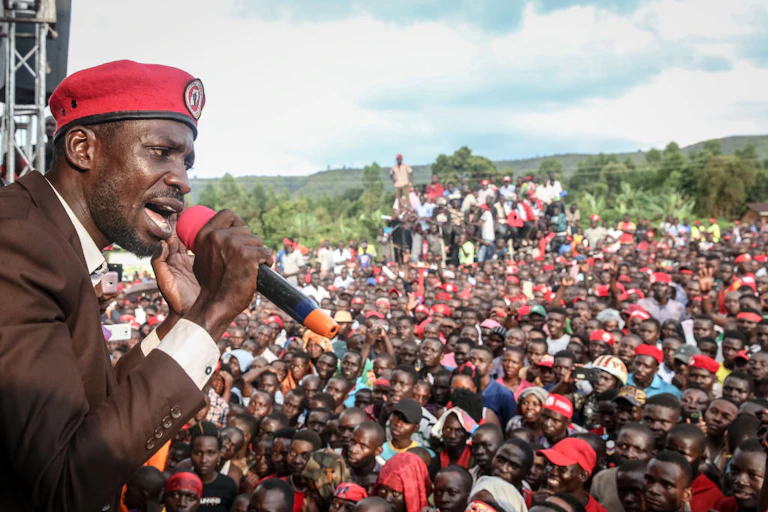 A man in a suit and red cap speaking into a microphone in front of a crowd of men and women, many of them in red