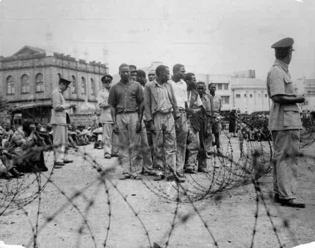 Black and white photo of men standing in a tight group and others sitting on the ground, watched by men in uniform, with barbed wire in the foregound.