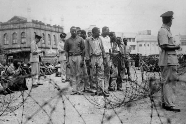 Black and white photo of men standing in a tight group and others sitting on the ground, watched by men in uniform, with barbed wire in the foregound.