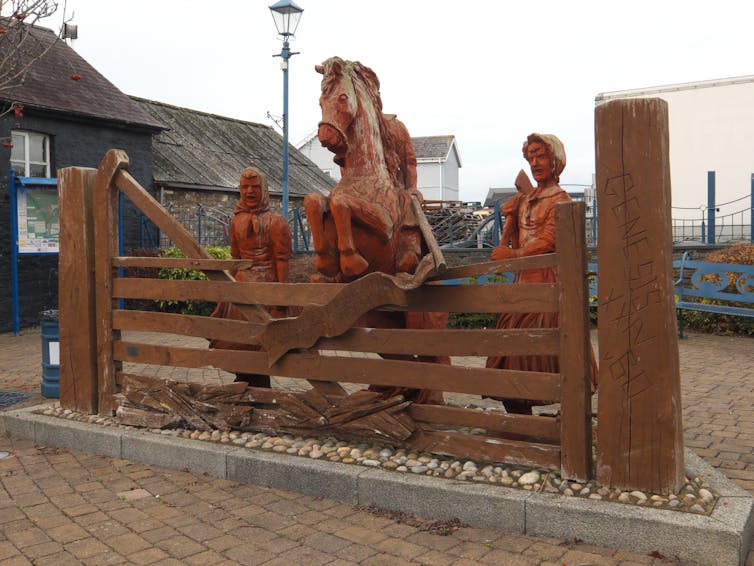 A wooden sculpture showing a horse flanked by two women leaping over a gate.