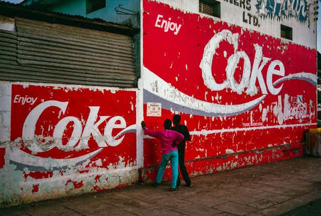 Two boys lean against a wall reading a poster. The wall is covered in a huge red and white mural advert reading Enjoy Coke.