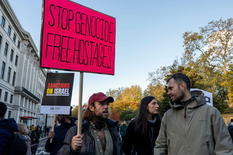 A man holds a sign saying Stop Genocide Free Hostages with another placard behind saying Sactions on Israel