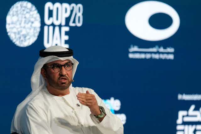 Un homme en thawb blanc s'exprimant sur un fond bleu avec "COP28 UAE" en lettres blanches.