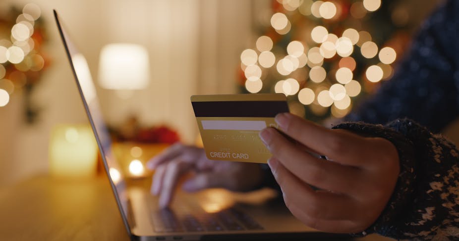 A close up of a hand holding a credit card with a laptop and Christmas lights in the background.