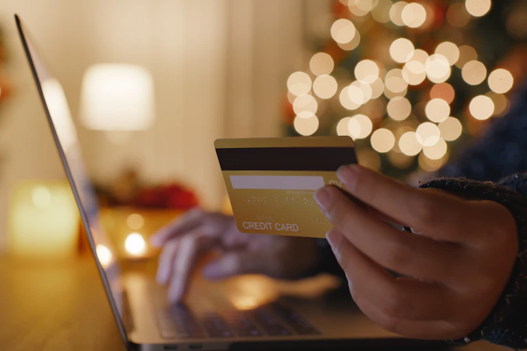 A close up of a hand holding a credit card with a laptop and Christmas lights in the background.
