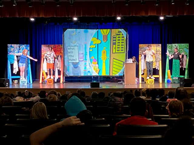 A stage in an auditorium with four people, each dressed in one color (from left to right blue, red, yellow and green), with a painting behind them of the same color. 