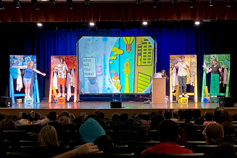 A stage in an auditorium with four people, each dressed in one color (from left to right blue, red, yellow and green), with a painting behind them of the same color.