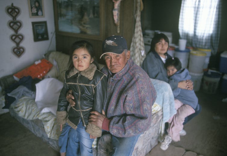 Native American grandfather sitting close to granddaughter in a bedroom, with a middle-aged woman and a child embracing in the background.