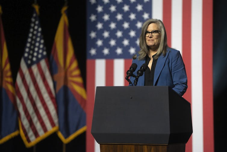 A white woman stands in front of an American flag as she delivers a speech.