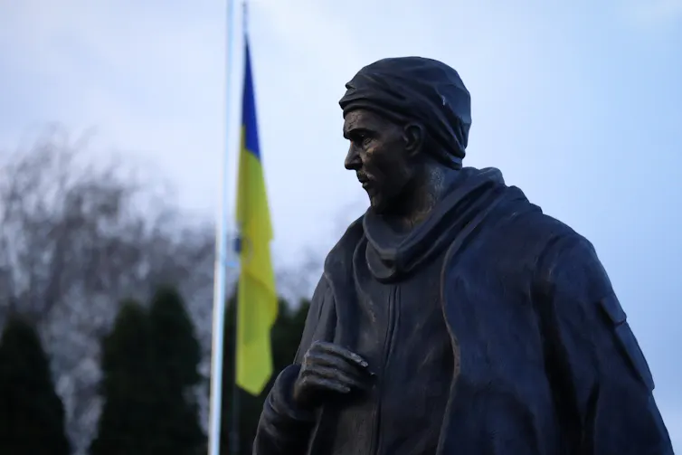 The statue of a soldier stands in front of a Ukrainian flag.