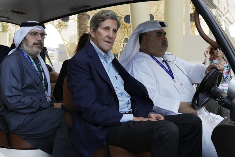 Kerry, in western business suit, rides in a golf cart outside the COP28 conference center with two men, both in traditional regional dress.