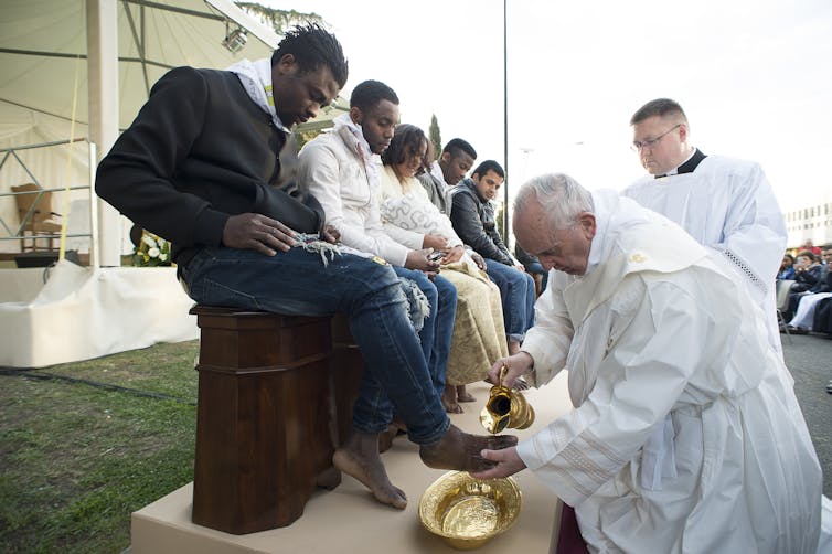 Several people seated in a row watch as the pope washes the feet of one of them.