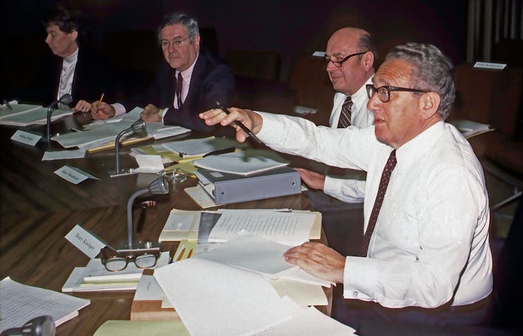 Un hombre sentado ante un escritorio da indicaciones a otros tres hombres que se encuentran a su lado