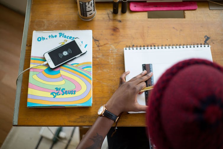 A student works at a desk with a notebook, ruler, phone and books.