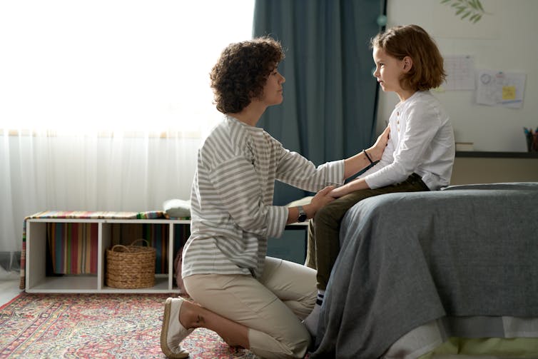 A mother talks to her child who is sitting on a bed.