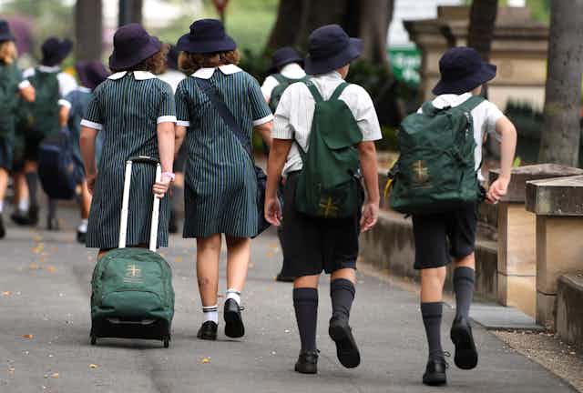 School students in matching uniforms, bags and hats walk along a street. 