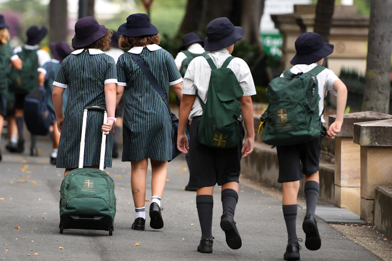 School students in matching uniforms, bags and hats walk along a street.