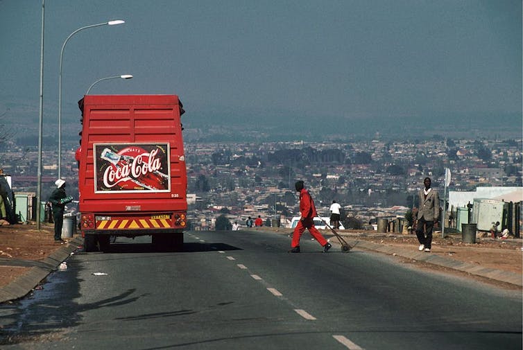 A red truck with the Cola-Cola logo on the back drives into a township dense with houses.