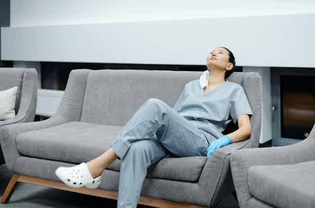 A tired woman doctor in scrubs and Crocs, reclining on a couch