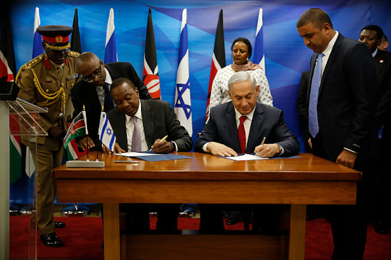 Two men seated at a table are holding pens over paper while others watch. There are flags in the background.