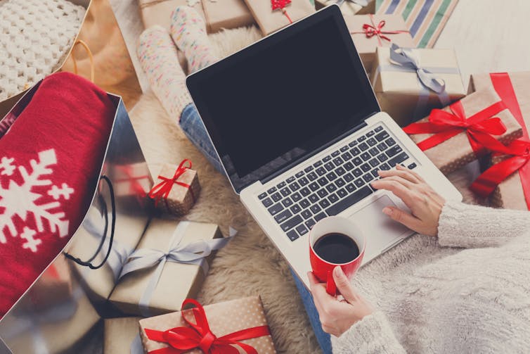 A person sits at a laptop with a coffee surrounded by festive packages.