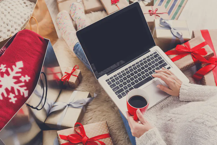 A person sits at a laptop with a coffee surrounded by festive packages.