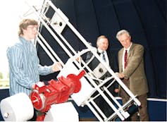 A telescope inside a dome during daytime, with a young teen and two older men standing next to it