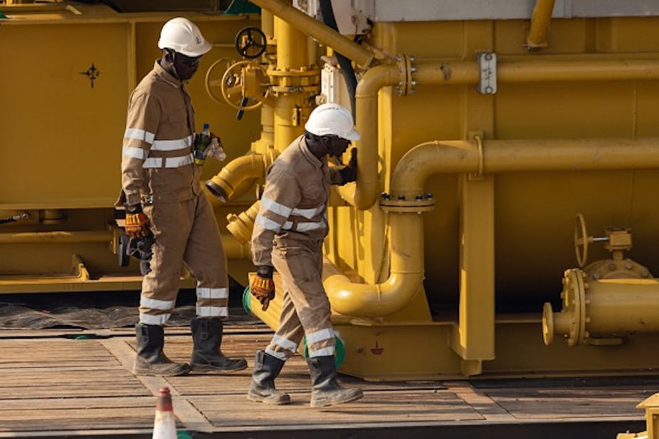 Two people in overalls, boots, gloves and hard hats look at large metal pipes painted yellow. 