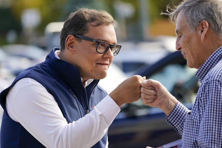 A brown haired man with glasses, wearing a white shirt and blue vest, fistbumps another man.