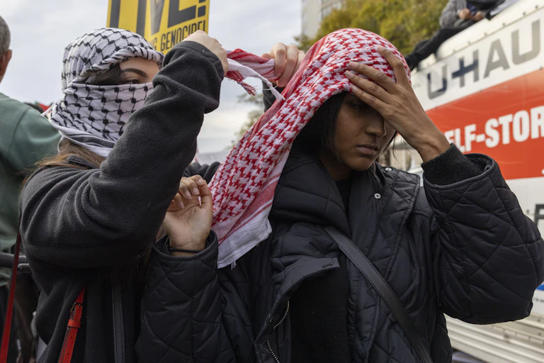 A person with a black checkered scarf wrapped around their head is tying a red checkered scarf around the head of another person.