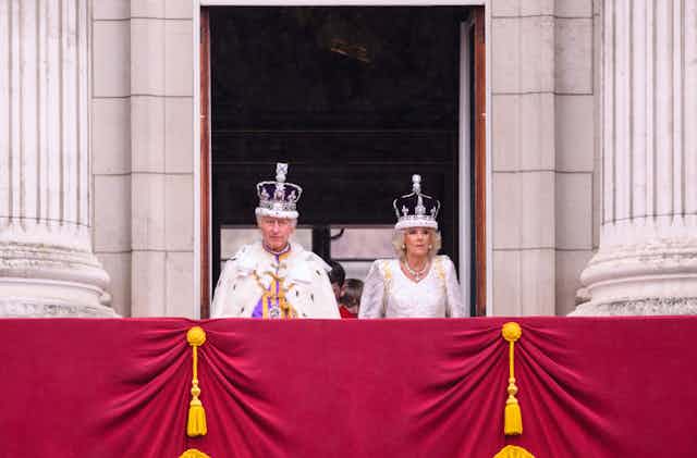 King Charles and Queen Camilla standing on a balcony in their crowns.