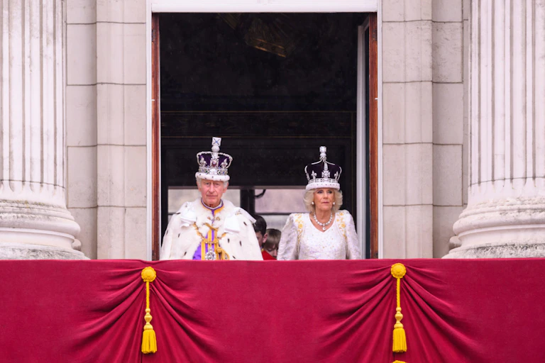 King Charles and Queen Camilla standing on a balcony in their crowns.