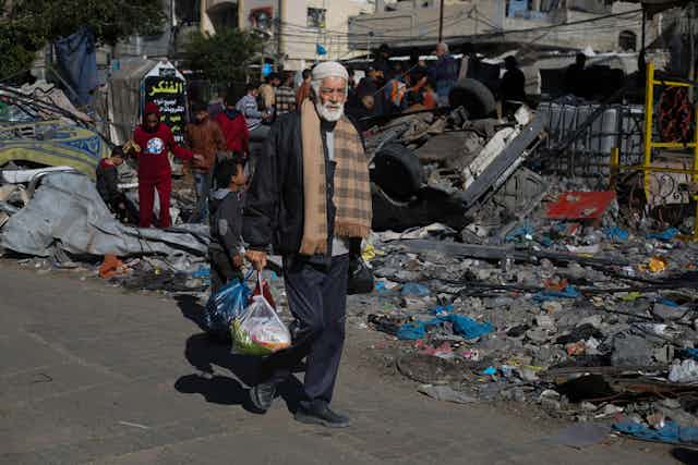 An elderly man walks down a street filled with rubble carrying food in carrier bags.