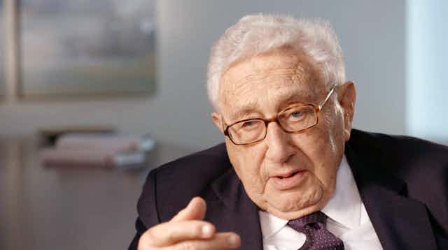 Henry Kissinger looks directly at the camera, wearing glasses, white shirt and red tie.