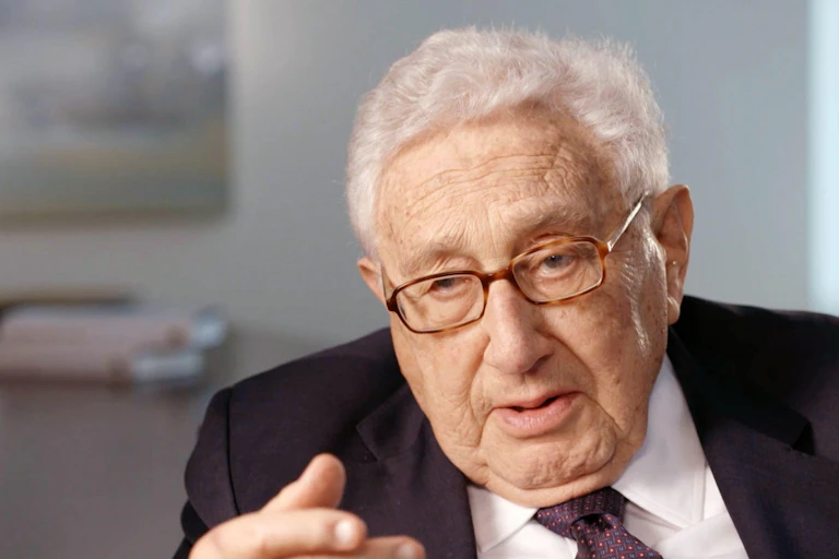 Henry Kissinger looks directly at the camera, wearing glasses, white shirt and red tie.