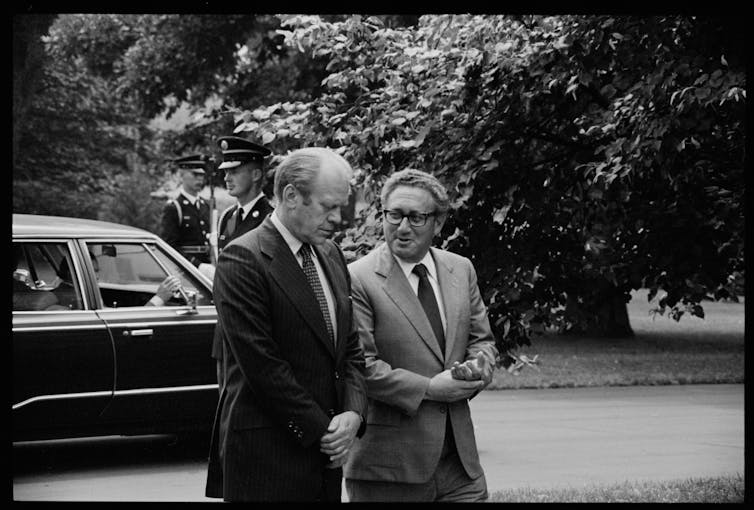 Two men walking with car in background, US president Gerald Ford and secretary of state Henry Kissinger, conversing, on the grounds of the White House
