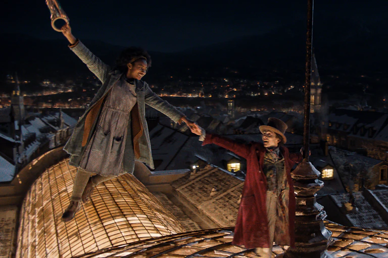 A smiling Black girl holds the hand of a white man wearing a top hat. They are on the roof of a building.