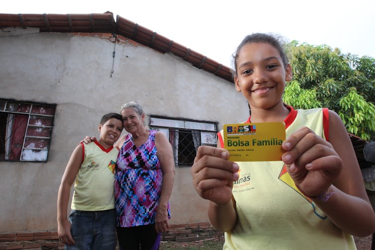 Family stand outside home, woman and boy in background smiling, girl in foreground smiling and holding a Bolsa Familia card.