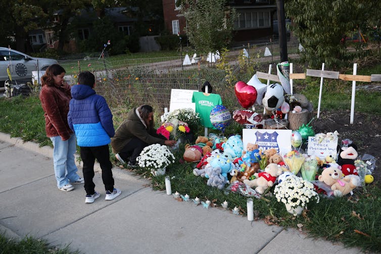 Three people stand and kneel next to a large pile of flowers, balloons and signs perched on grass, off of a sidewalk.